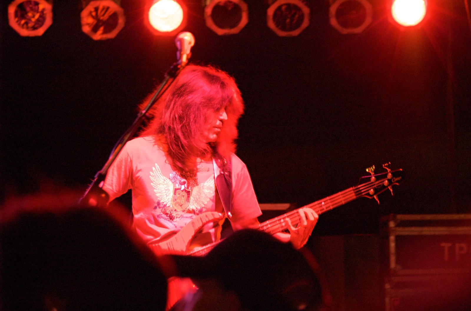 Rudy Sarzo playing bass guitar on a red-lit stage during a Blue Oyster Cult performance