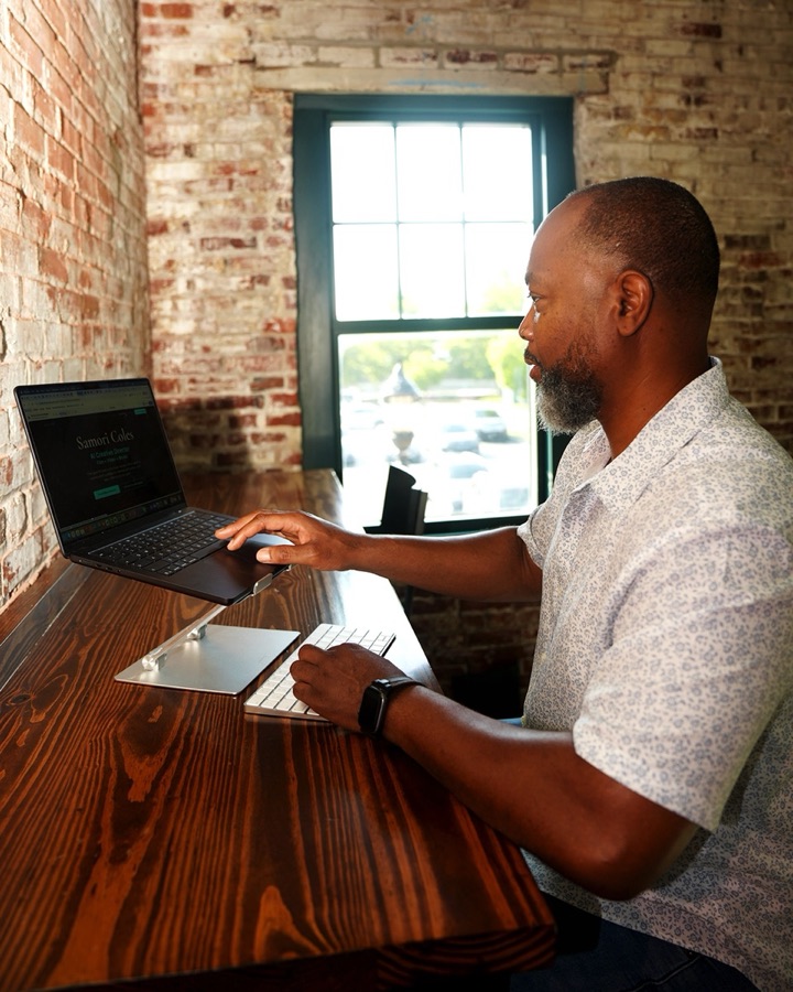 Samori Coles working at a laptop showing his AI creative director site, in a brick-walled room