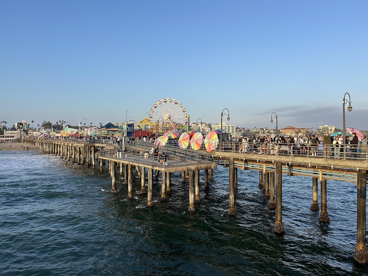 Crowds walking along Santa Monica Pier on a clear day, with colourful parasols, the Ferris wheel and lamp posts along the boardwalk.
