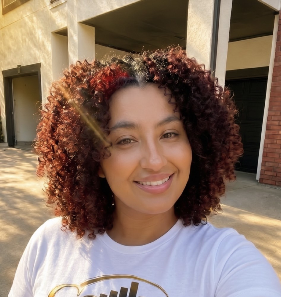 Sheena Taylor smiling outdoors in sunlight with reddish-brown curly hair and a white logo T-shirt.