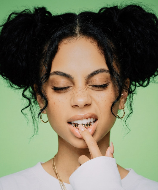 Slime Dot — studio portrait with double space buns, freckles, and a finger to her lips revealing a metal grill, against a mint-green backdrop.