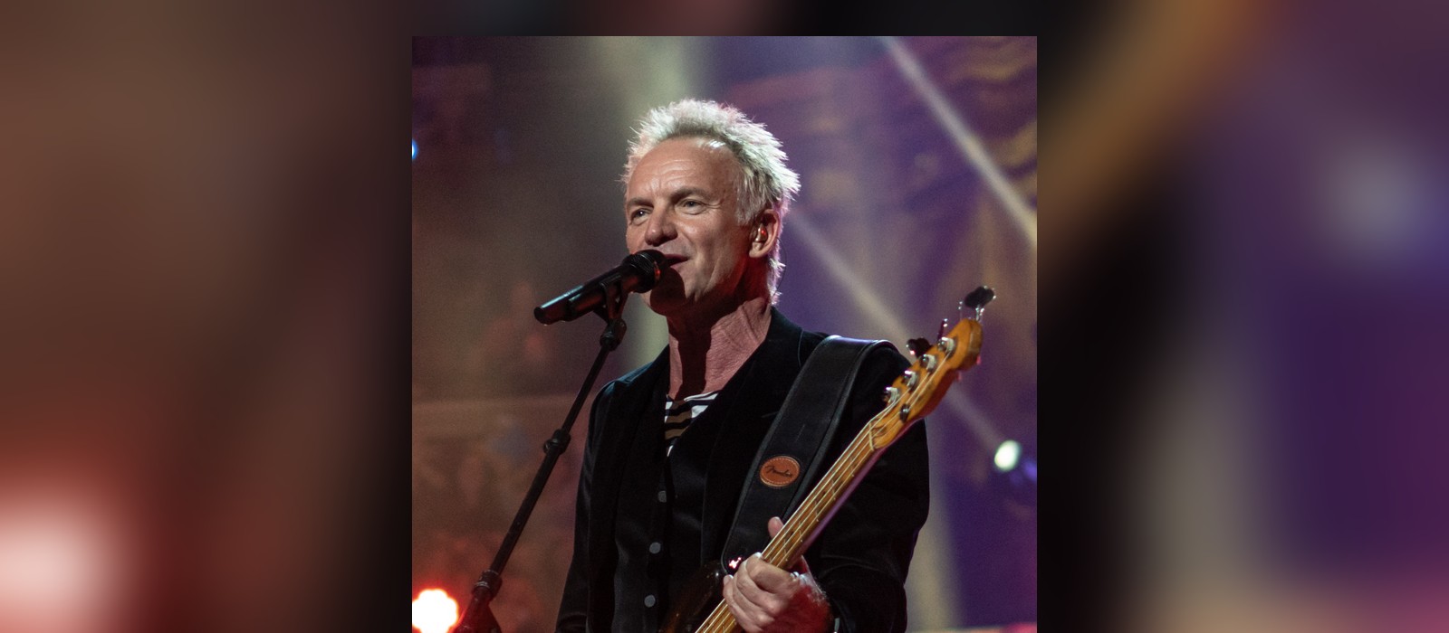 Sting singing into a microphone while playing bass guitar on stage, photographed in April 2018