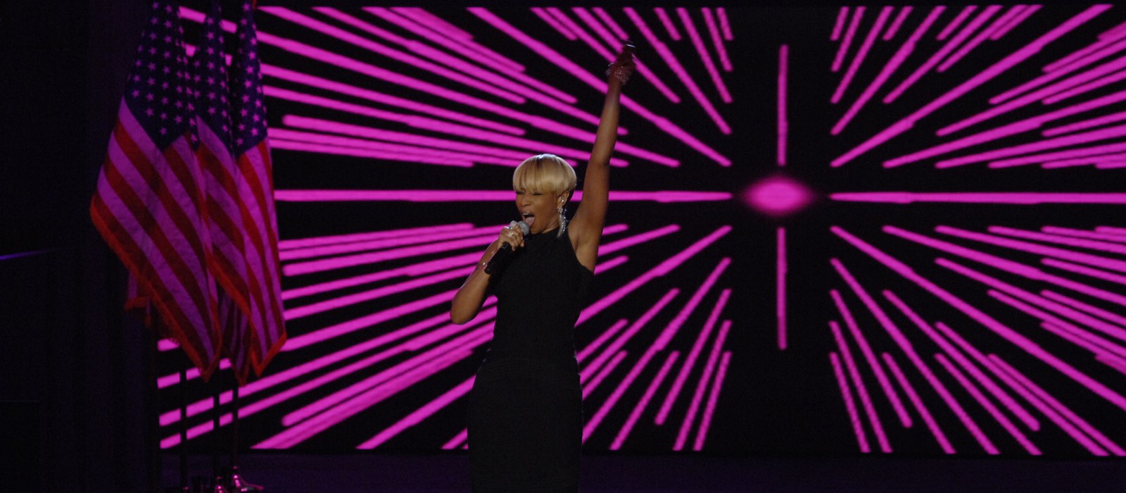Mary J. Blige performing on stage with one arm raised in front of a magenta starburst screen