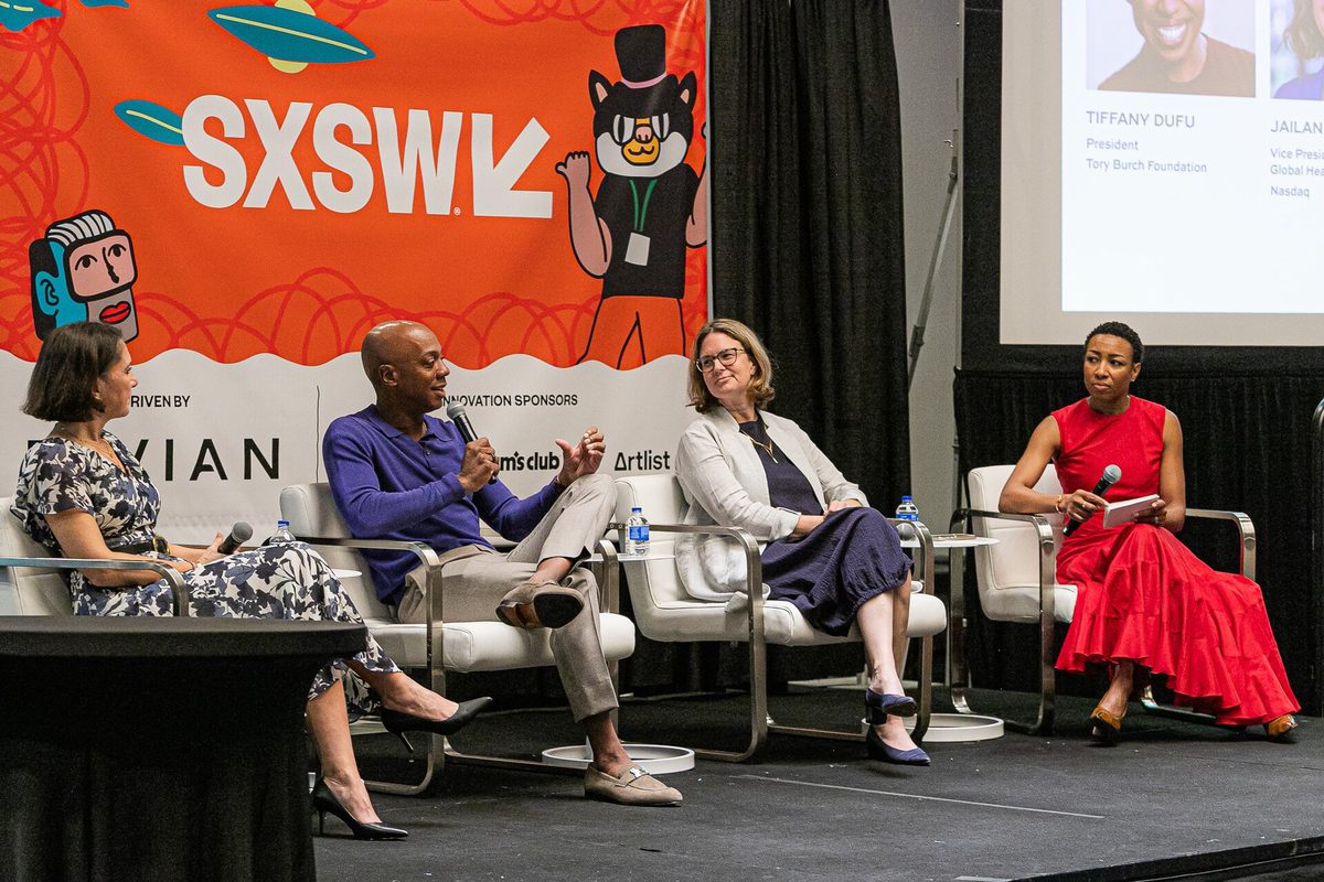 Four panellists on a SXSW stage against an orange SXSW backdrop, one speaking into a handheld microphone, with speaker headshots projected on a screen behind.