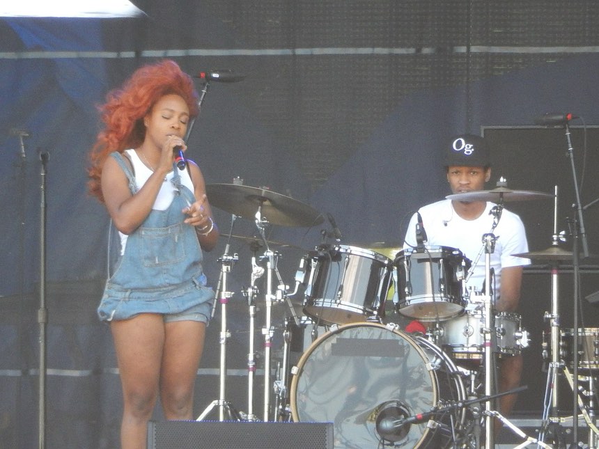 Singer SZA performing live on stage at Lollapalooza 2015, holding a microphone under concert lighting