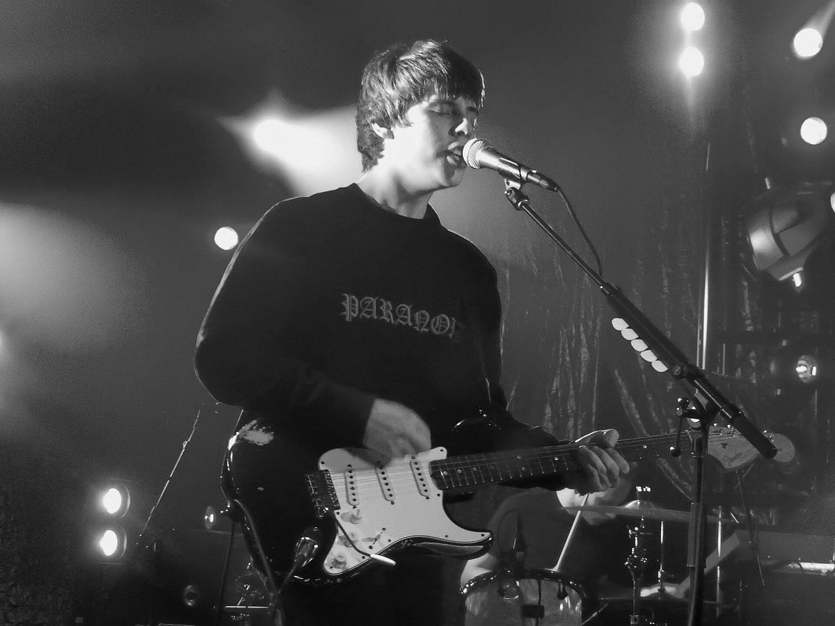 Jake Bugg singing into a microphone and playing a Fender electric guitar under white stage lights, shot in black and white.