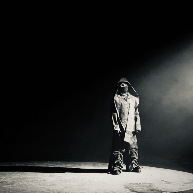 A hooded figure in a balaclava standing alone in a spotlight on a bare floor, shot in high-contrast black and white, face fully concealed.