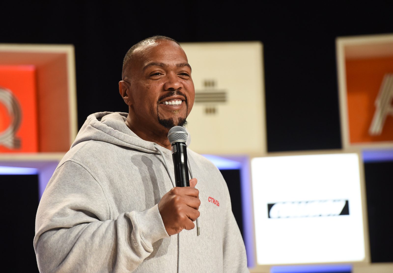 Timbaland smiling into a microphone on a conference stage in a grey hoodie