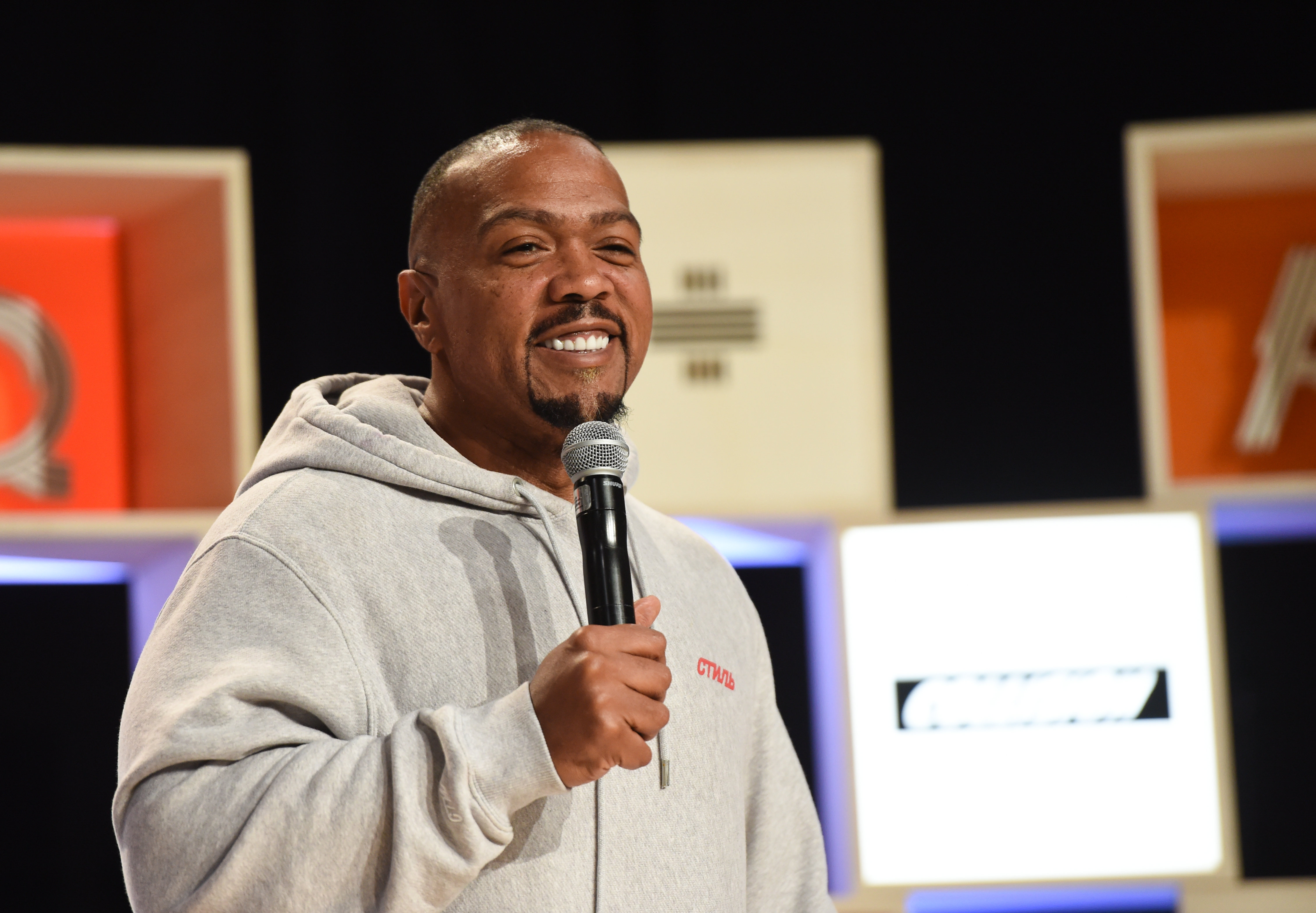 Timbaland smiling and speaking into a handheld microphone on stage in a grey hooded sweatshirt, in front of a dark backdrop with colourful geometric panels.