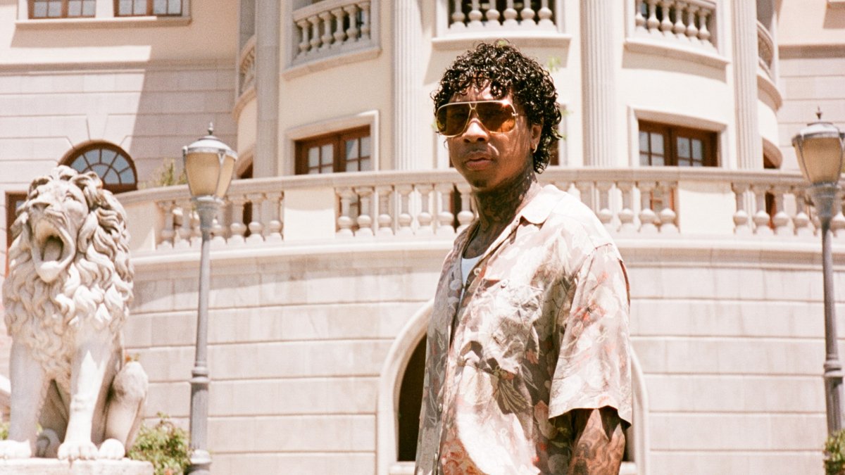 Tyga in sunglasses and a patterned shirt standing in front of a mansion, from his $TARFACE album rollout