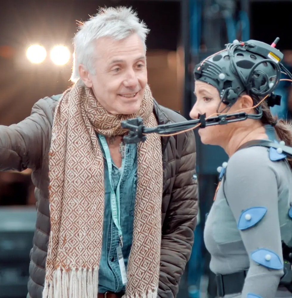 A man in a scarf talking to a performer wearing a motion-capture suit and head rig during a Unit1 Studio capture session