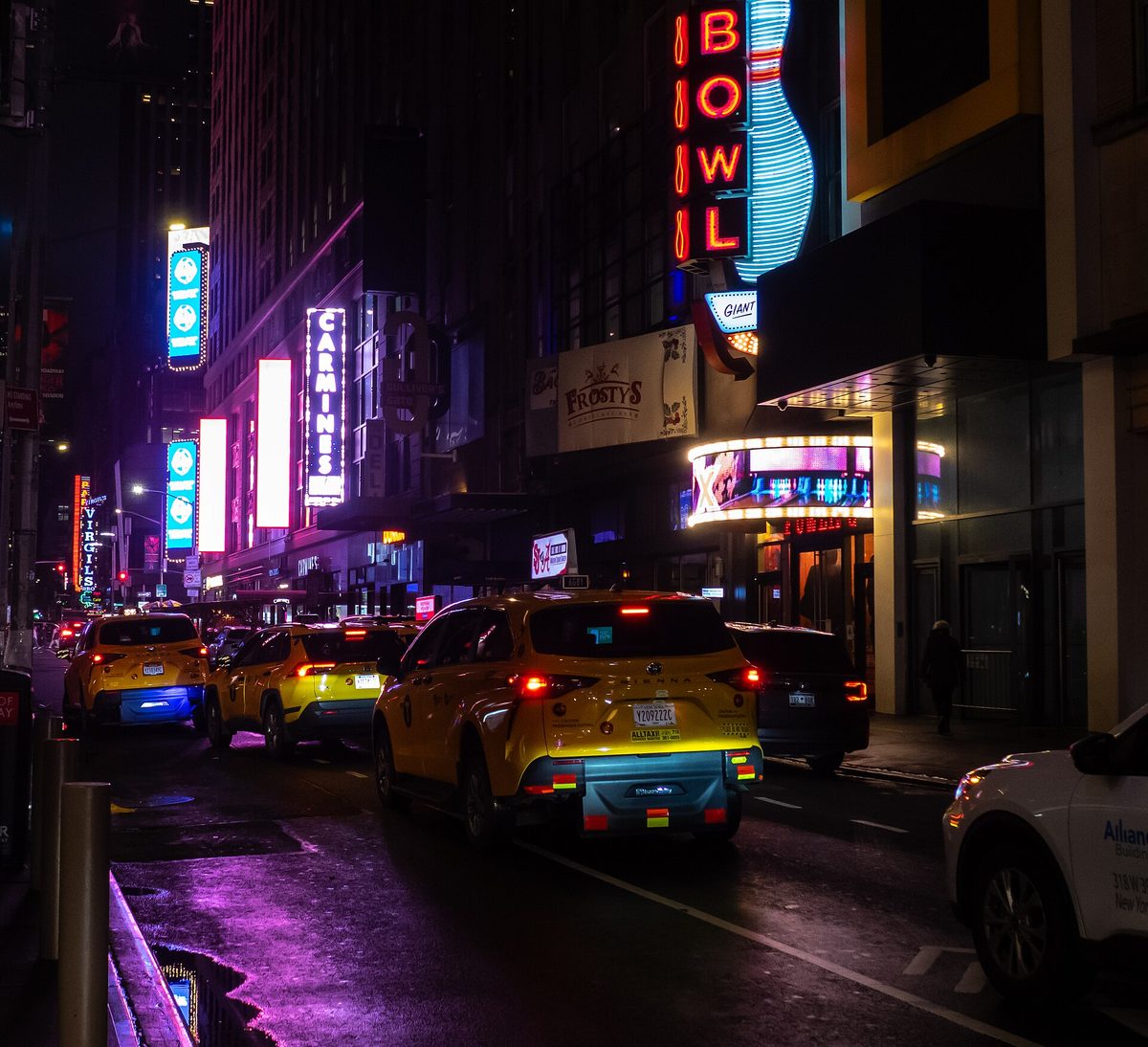 West 44th Street in New York at night, lined with neon bar and bowling signs, yellow taxis queueing on the wet road.