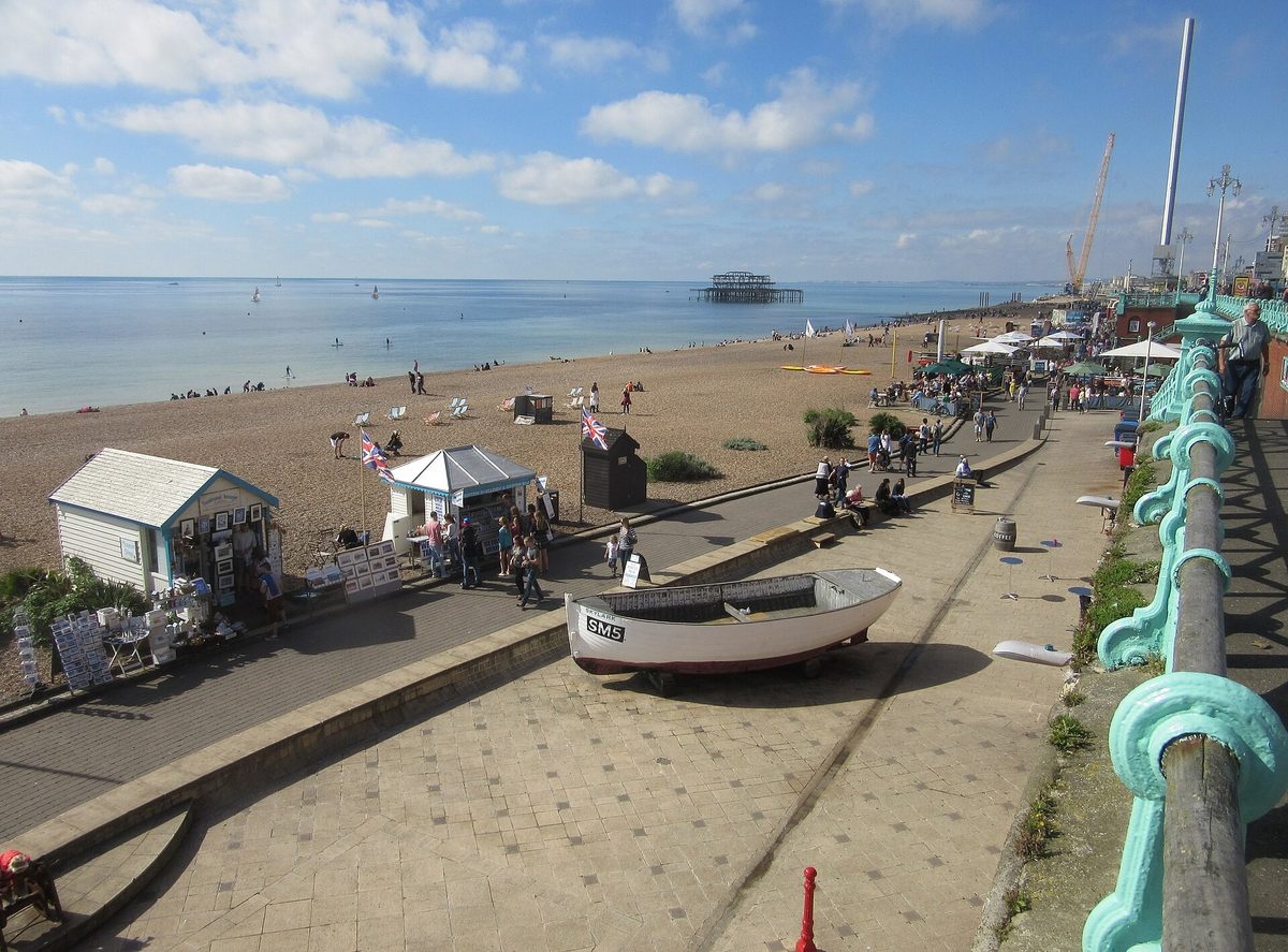 Brighton seafront looking west, with beach huts and stalls along the promenade and the burnt-out remains of the West Pier standing in the sea.