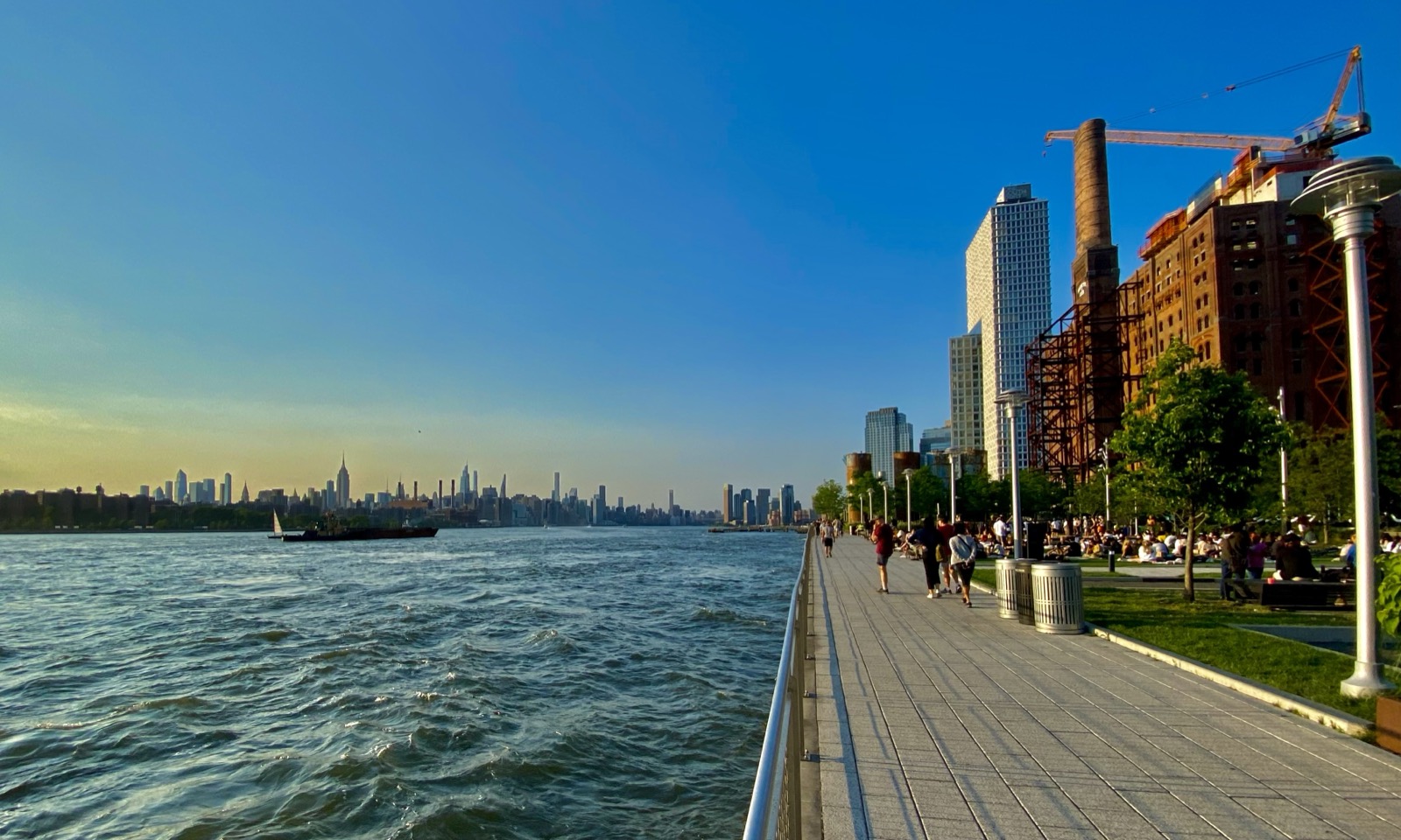 The Williamsburg waterfront promenade at Domino Park looking across the East River to the Manhattan skyline, with the converted Domino Sugar refinery on the right.