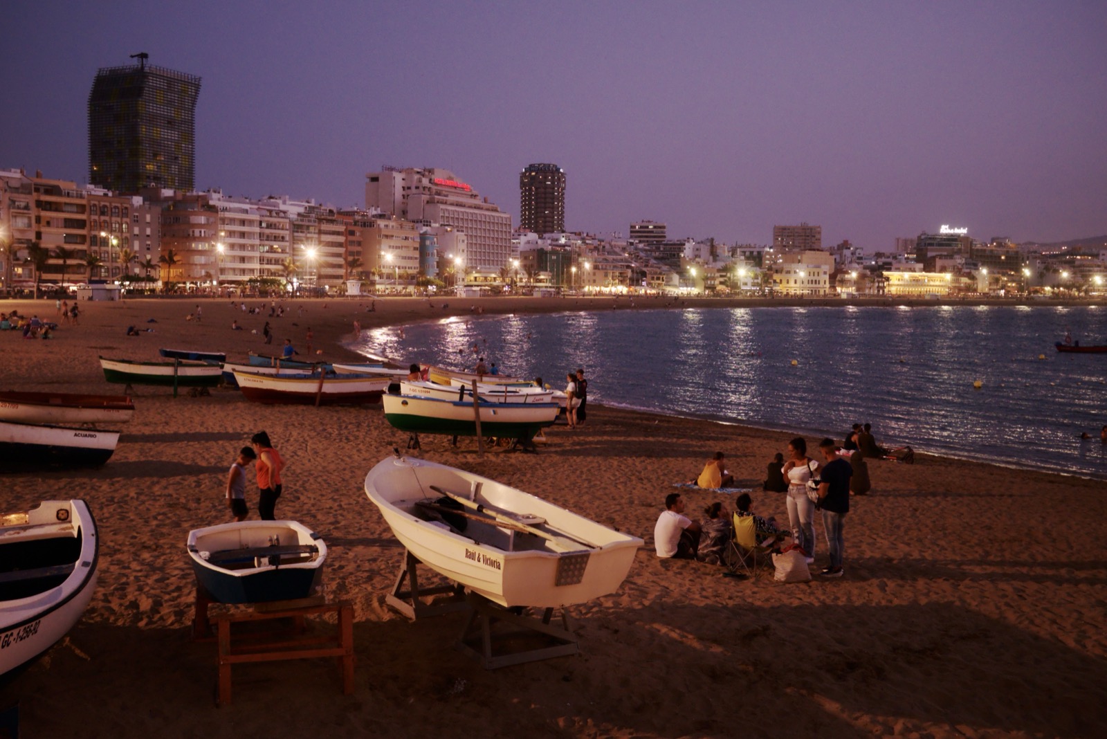 Playa de las Canteras in Las Palmas de Gran Canaria at dusk, with fishing boats pulled up on the sand and the lit city skyline curving around the bay.