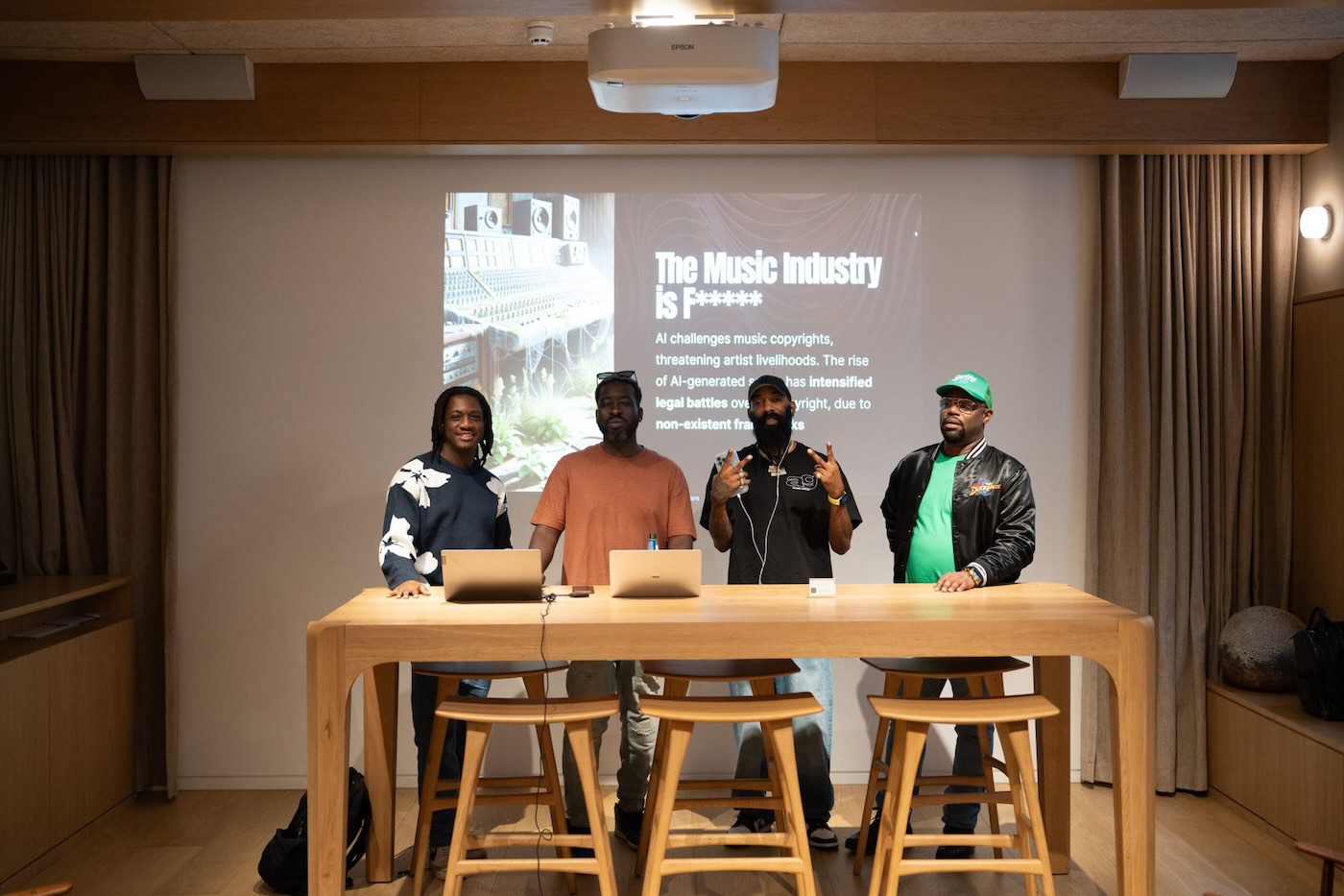 Twoshot panel of four men behind a long wooden table with laptops in front of a Music Industry slide