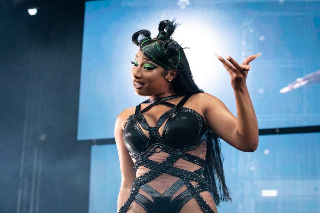 Megan Thee Stallion on stage wearing a black, strappy, and shiny bodysuit with intricate details, featuring cutouts and sheer panels. She has long, straight black hair with green highlights styled in two top buns and is wearing dramatic green eye makeup. She is gesturing with one hand while looking to the side, with a confident expression. The background shows a large, blurry screen and stage lights, creating a vibrant concert atmosphere.