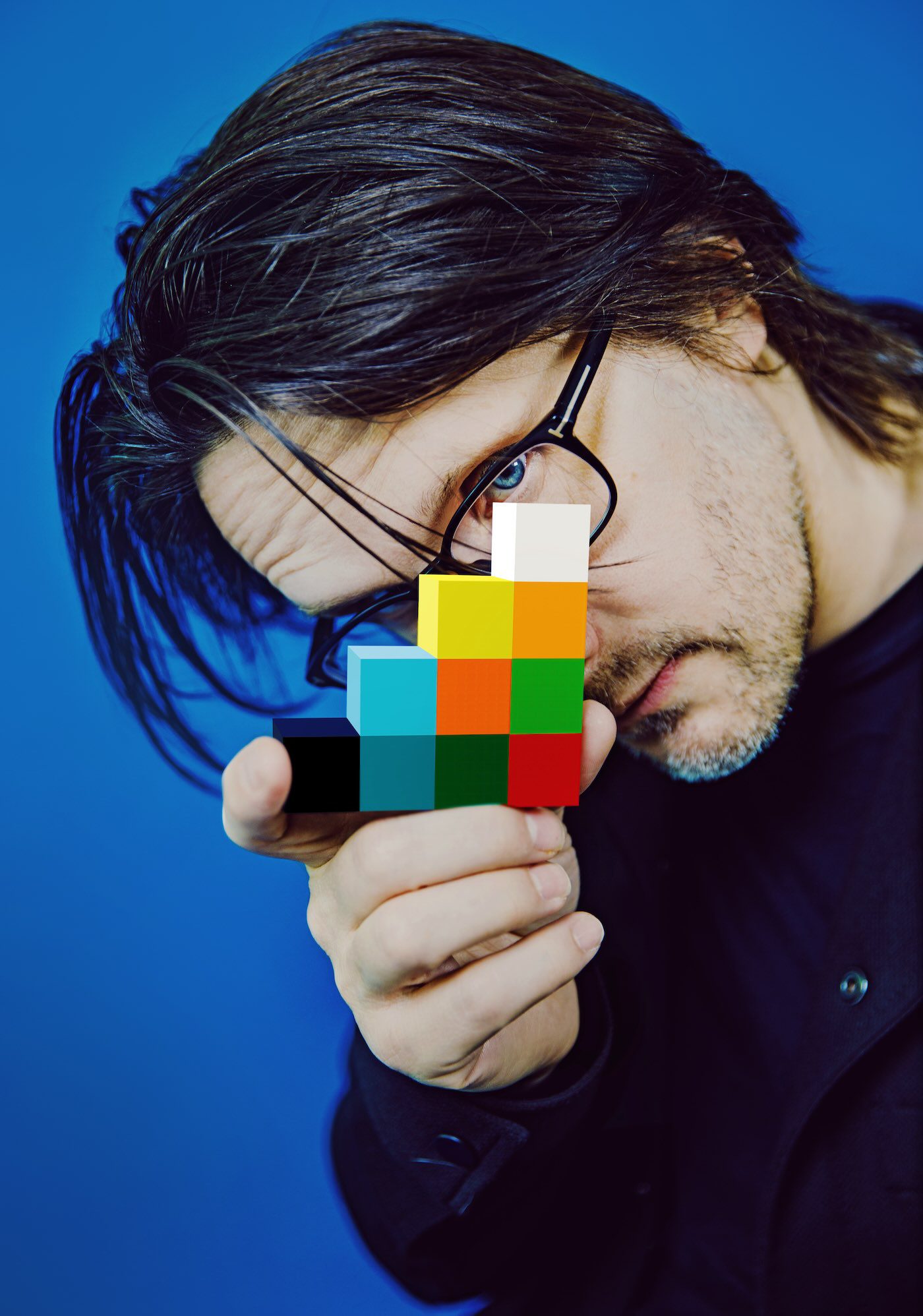 Steven Wilson with Blocks (Blue) – Steven Wilson peers over colorful stacked blocks against a blue backdrop.