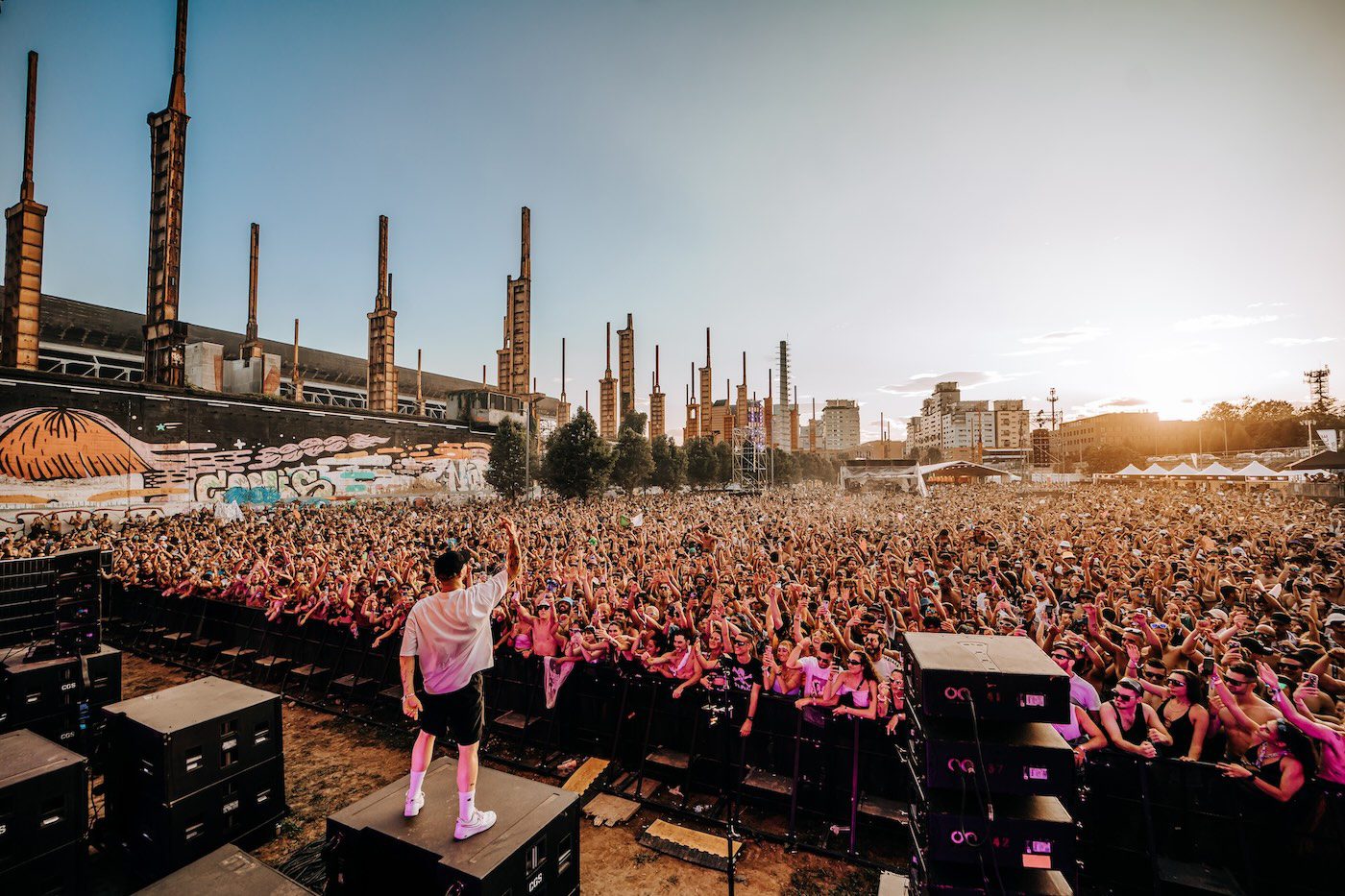 DJ on stage facing a massive crowd at Kappa Futur Festival with industrial cooling towers in the background at sunset
