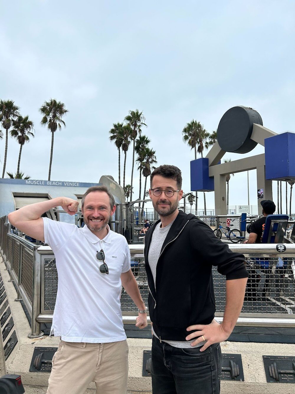 Two men posing in front of Muscle Beach Venice sign with palm trees behind them, one flexing in a white polo and the other in a black hoodie