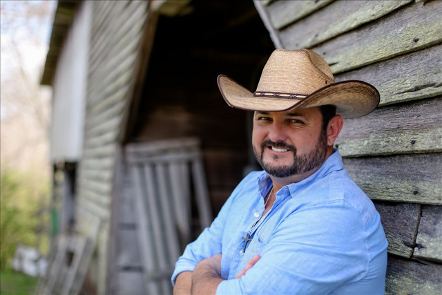 Tony Justice poses in a cowboy hat against a rustic barn, representing his country artist brand.