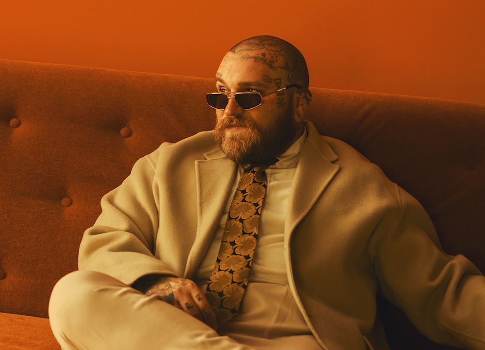 Teddy Swims sitting on an orange couch in a beige suit and patterned tie, studio photo session.