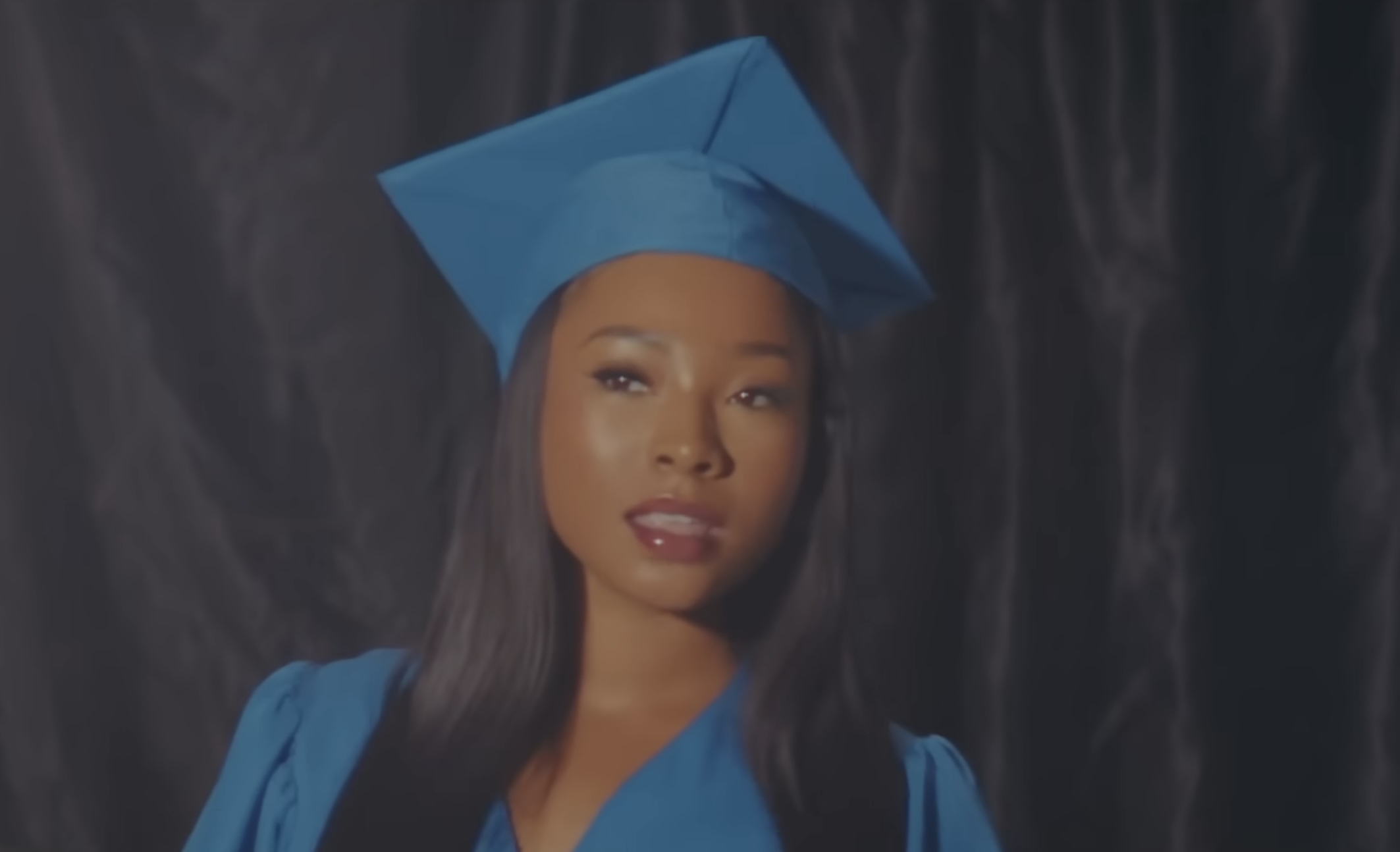 Actress poses confidently in a blue graduation cap and gown in the "How Was I Supposed to Know" music video by Xania Monet.