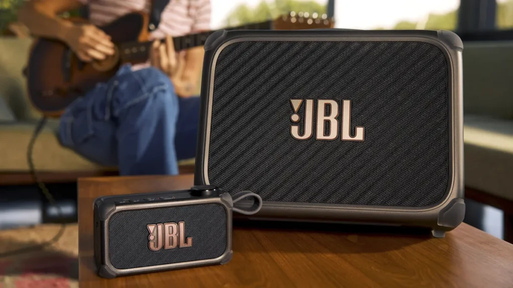 Two JBL speakers (Bandbox) on wooden table with person holding guitar in background.