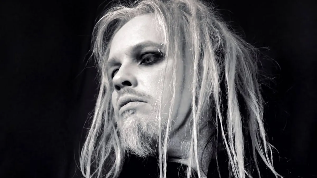 Darkwave Musician Anders Manga with long blonde hair and dramatic eye makeup looks upward against dark background.