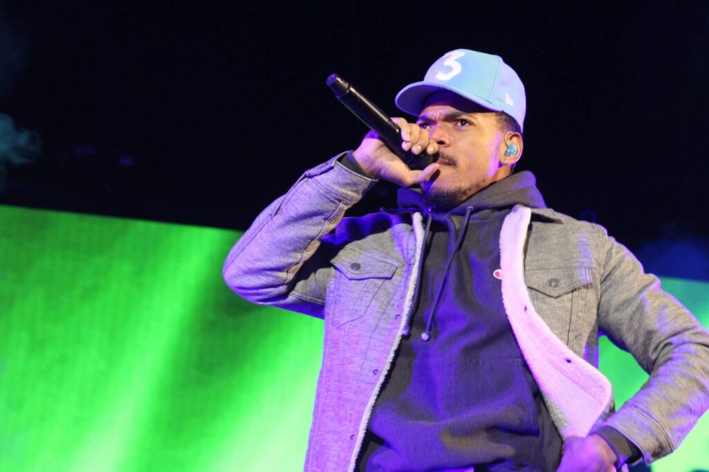 Chance the Rapper performing at Red Rocks, wearing signature '3' cap, holding microphone on stage.