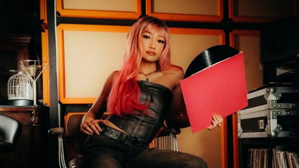 TaTa Taktumi holding a vinyl record in a music studio surrounded by record crates and vintage gear.