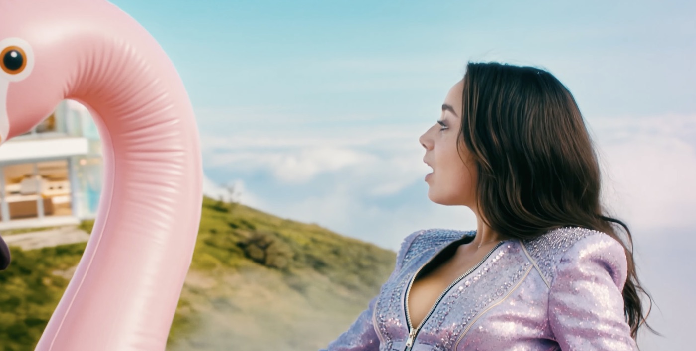 Tilly Norwood in a sparkly lavender jacket gazing at clouds above a hillside, with a giant pink flamingo float beside her.