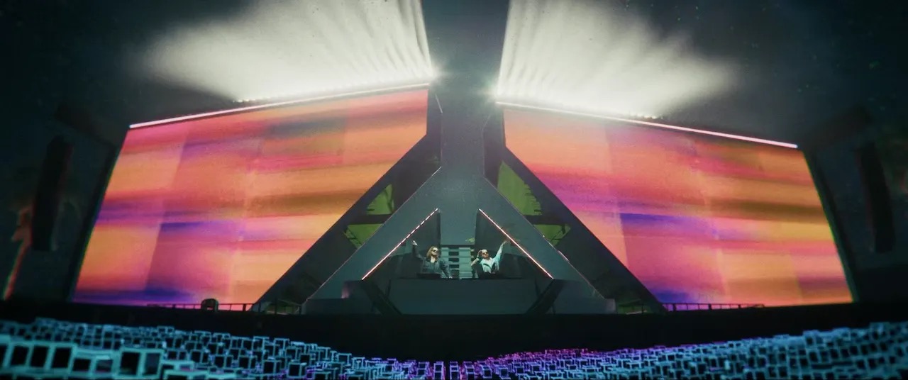 Two DJs perform at a futuristic triangular stage with huge rainbow LED screens behind them.