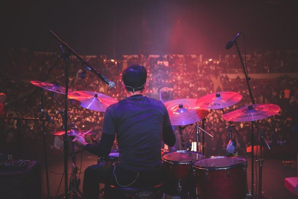 Drummer viewed from behind playing live on stage in front of a packed arena crowd with red cymbals.