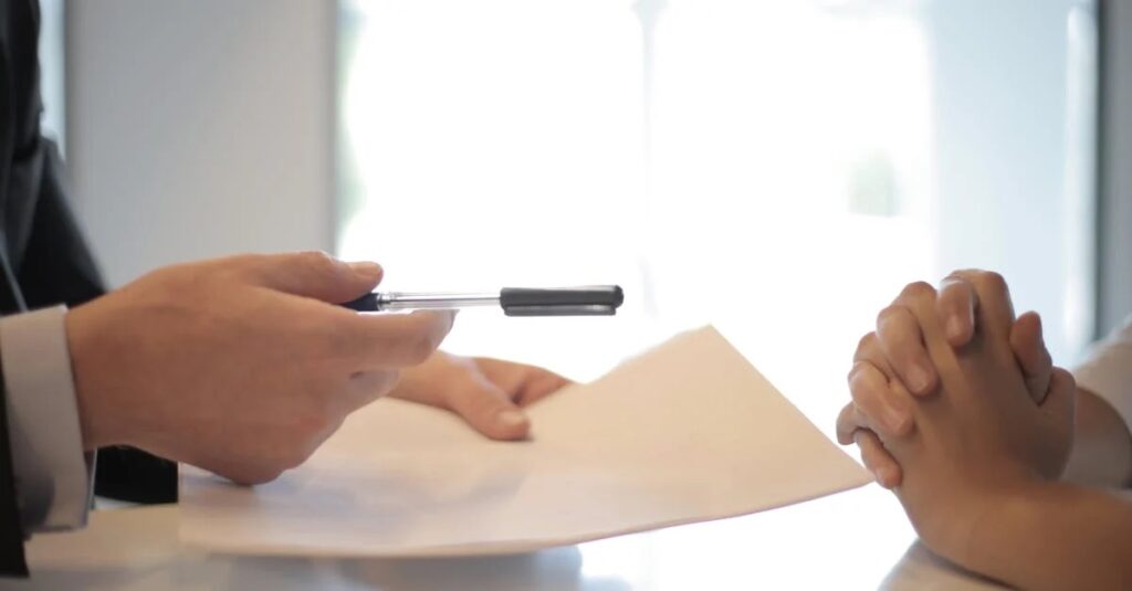 Two people at a desk with one person handing over a pen and contract document for signing in a business meeting.